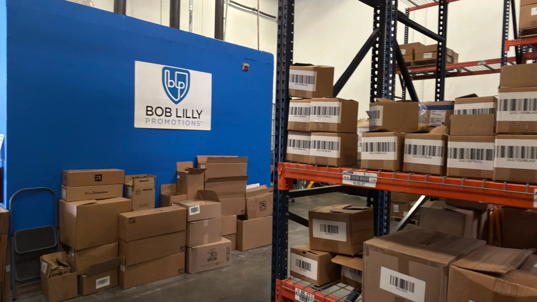 Branded gift cartons staged for shipment beside the Bob Lilly Promotions logo wall in our Garland, Texas fulfillment center
