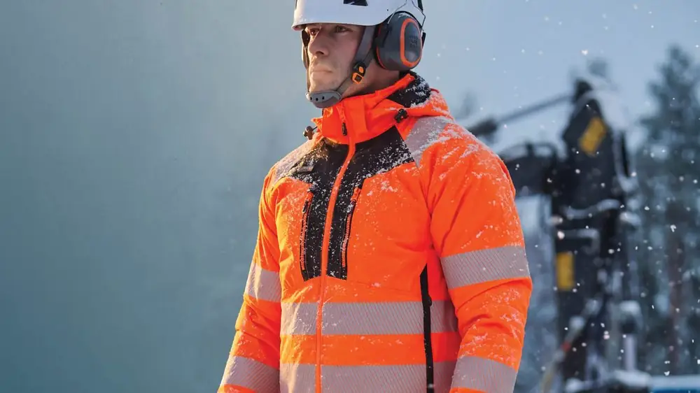 Worker in an orange hi-vis insulated jacket, safety helmet and ear protection in snow
