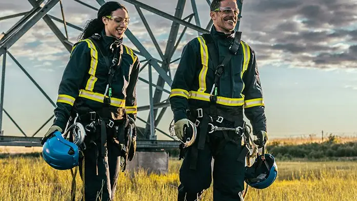 Two workers in hi-vis reflective workwear and fall protection harnesses, holding safety helmets